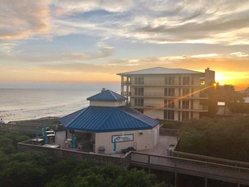 A beachfront view at sunset, featuring a round building with a blue roof and a multi-story building in the background.