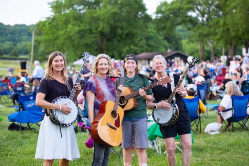 Four musicians stand together in a park, each holding instruments, with a crowd enjoying a concert in the background.