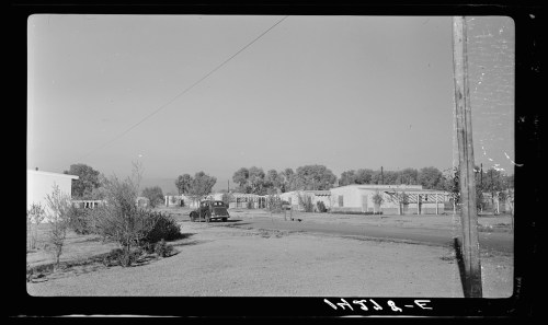 A vintage car parked on a dirt road near small buildings and trees in a rural setting.