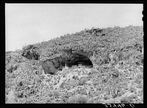 A rocky hillside with a cave entrance, surrounded by desert vegetation and cacti.