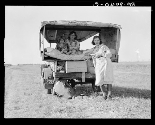 A family of four sits in a truck bed, with a woman standing outside, in a rural setting.