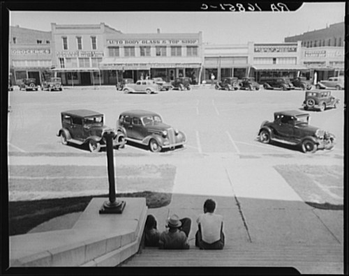 A historic street scene featuring vintage cars parked in front of shops, with three people sitting on steps.