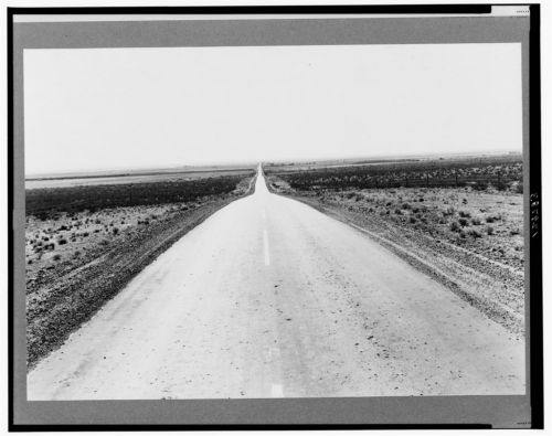 A long, straight road stretches into the horizon, surrounded by barren land and sparse vegetation. Black and white image.