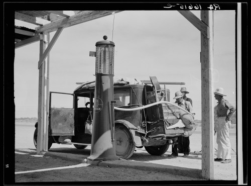 A vintage car at a gas station with two men working on it, under a wooden structure in a desert setting.