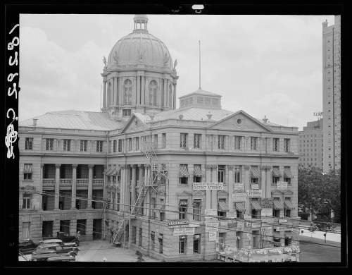 Historic building with a large dome, featuring banners and a fire escape, surrounded by trees and cityscape.