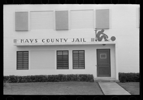 Black and white image of Hays County Jail exterior, featuring a sign and a decorative figure playing a horn.