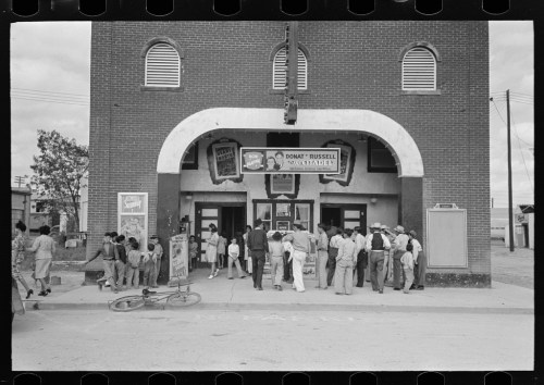 A crowd gathers outside a vintage theater with a marquee, featuring a film poster and a bicycle nearby.
