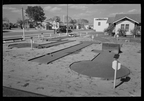 A black and white photo of a mini-golf course with several holes and obstacles, surrounded by a sandy area.