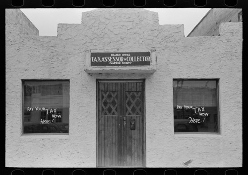 Front view of a tax assessor and collector office with a textured wall and windows displaying "Pay Your Tax Now Here!" signs.