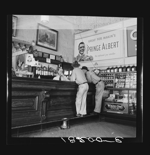 Two men converse at a bar counter, with a sign for "Prince Albert" tobacco in the background. Vintage setting.