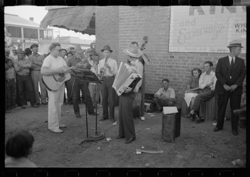 A group of musicians perform outdoors, with a guitarist, accordionist, and others playing instruments in a lively setting.
