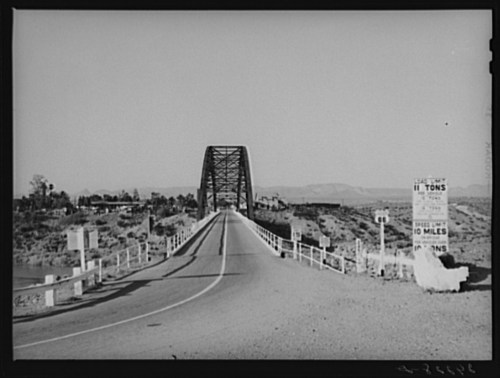 A black and white photo of a bridge over a river, with signs indicating weight limits and distance.