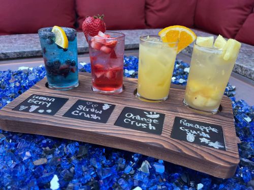 Four colorful cocktails on a wooden tray, labeled: Lemon Berry, Straw Crush, Orange Crush, and Pineapple Crush.
