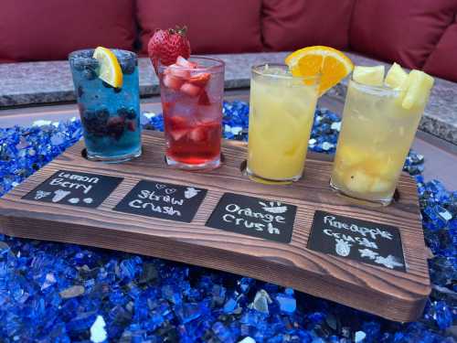 Four colorful cocktails on a wooden tray, labeled: Lemon Berry, Straw Crush, Orange Crush, and Pineapple Crush.