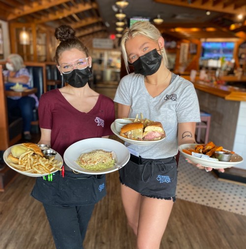 Two waitstaff in masks hold plates of food, including fries and sandwiches, in a restaurant setting.