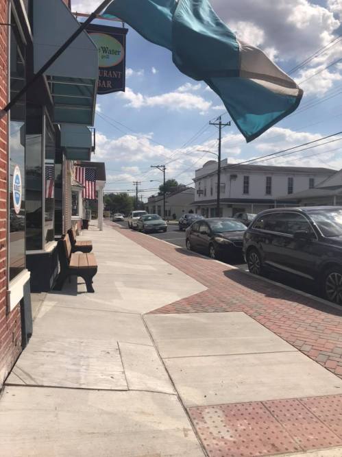 A street view featuring a sidewalk, parked cars, and a restaurant sign under a blue sky with clouds.