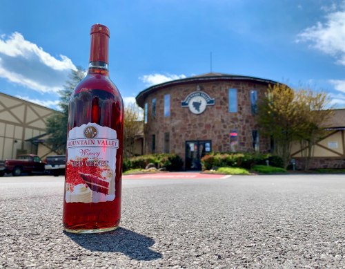 A bottle of Mountain Valley Winery wine in front of a stone building under a blue sky with scattered clouds.