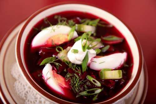 A bowl of borscht topped with sour cream, sliced eggs, green onions, and fresh herbs.