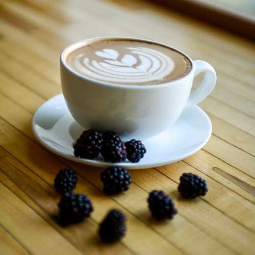 A cup of latte art on a white saucer, surrounded by fresh blackberries on a wooden table.