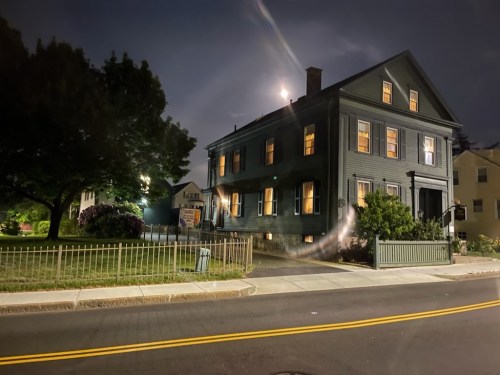 A dark blue house at night, illuminated windows, surrounded by trees and a fence, with a full moon in the background.