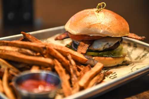 A juicy burger with bacon, lettuce, and tomato, served with crispy fries and a side of ketchup on a metal tray.