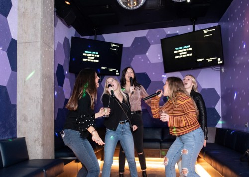 A group of five women singing karaoke in a vibrant, colorful room with screens and disco lights.