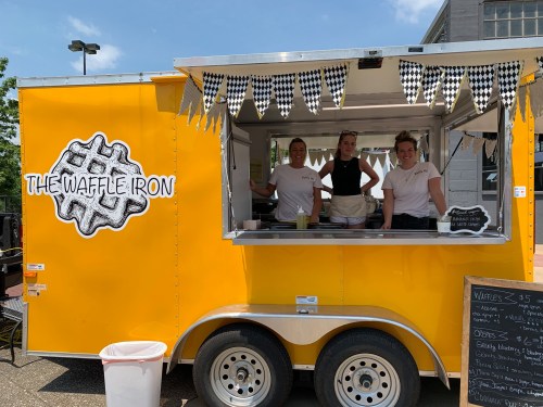 A bright yellow food truck named "The Waffle Iron" with three smiling staff members serving waffles.