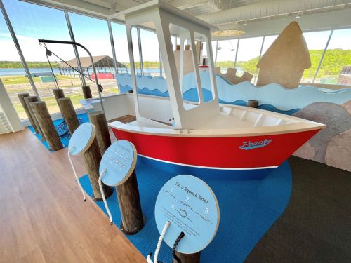 A colorful children's play area featuring a red boat and interactive displays, with a scenic view of water and hills outside.
