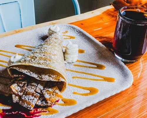 A plate with a crepe cone filled with sweet toppings, drizzled with sauce, and a glass of dark beverage beside it.