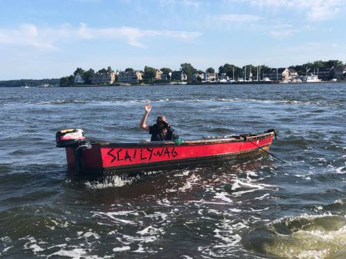 A person waves from a small red boat named "Scallywag" on a calm waterway with houses in the background.