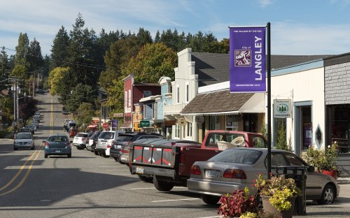 A quaint street in Langley, lined with shops and parked cars, surrounded by trees and a clear blue sky.
