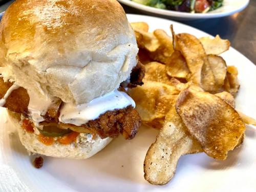 A crispy chicken sandwich with sauce and pickles, served with a side of potato chips on a white plate.