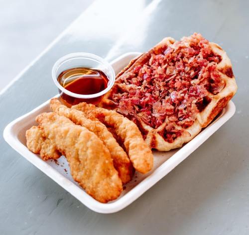 A plate featuring crispy chicken tenders, a bacon-topped waffle, and a small cup of syrup.