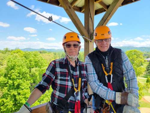 Two people wearing helmets and harnesses stand on a platform, surrounded by trees and mountains under a blue sky.