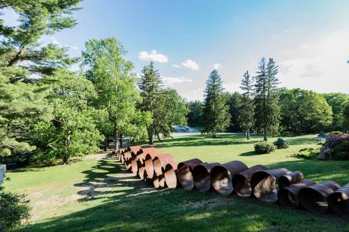 A row of rusty metal pipes lies on a grassy landscape, surrounded by trees and a clear blue sky.