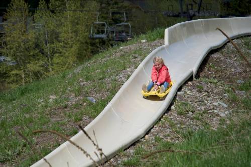A child in a red jacket slides down a smooth, curved slide on a grassy hill, surrounded by trees.
