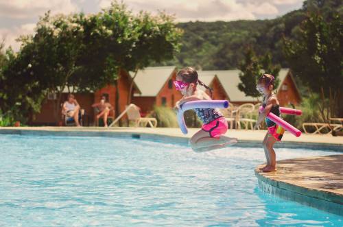 Two children jump into a pool with pool noodles, while adults relax nearby in a sunny outdoor setting.