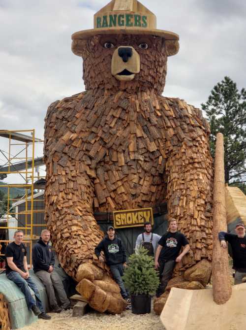 A large wooden bear sculpture with a ranger hat, surrounded by five people and a small tree.