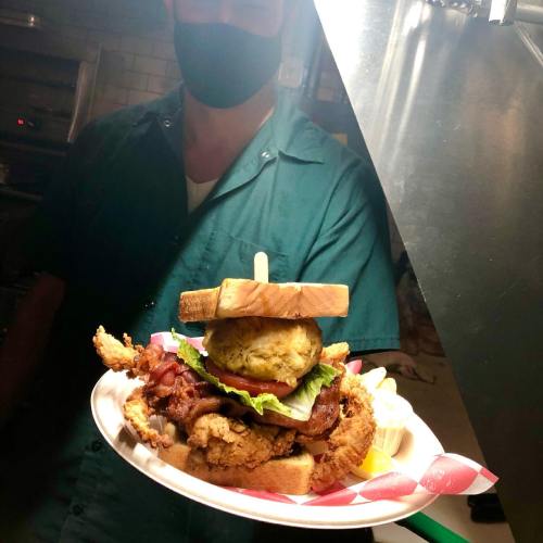 A person in a mask holds a large sandwich with fried chicken, bacon, lettuce, and two slices of bread on a plate.