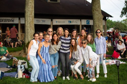 A group of women smiling and posing together outdoors, with a building and people in the background.