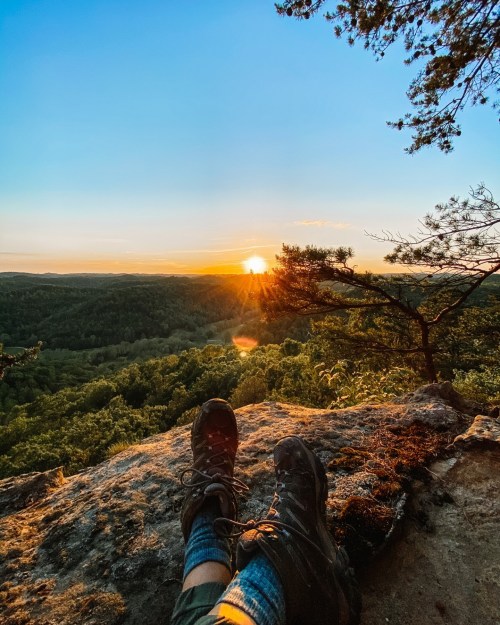 A pair of hiking boots resting on a rocky ledge, overlooking a sunset over a lush green landscape.