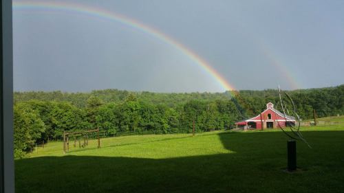 A vibrant rainbow arcs over a green landscape with a red barn in the distance, under a cloudy sky.