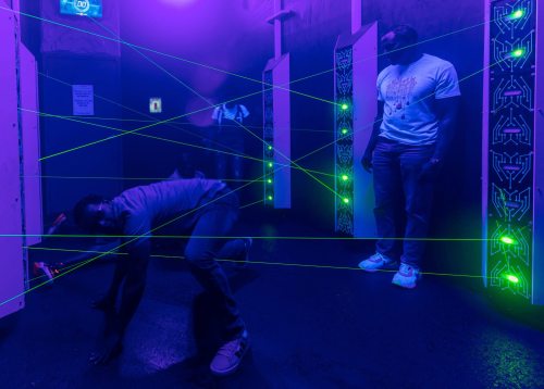 A person crawls under green laser beams in a dimly lit room, while another watches nearby.