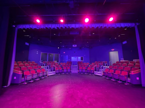 A theater stage with purple lighting, featuring red seats arranged in a semi-circle and a dark floor.
