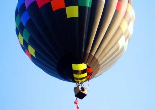 A colorful hot air balloon floats against a clear blue sky, with a person in the basket and a flag hanging below.