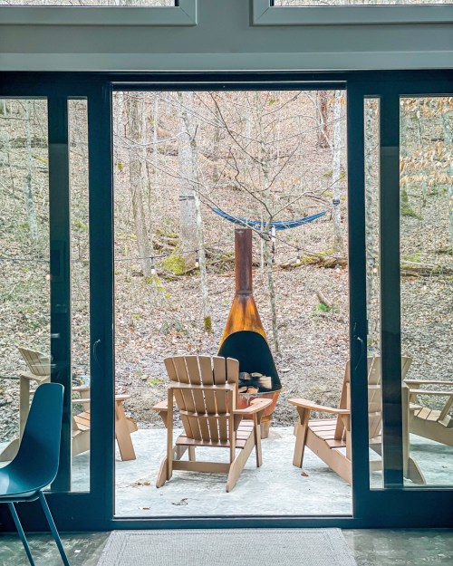 View through large glass doors to a serene outdoor space with wooden chairs and a fire pit surrounded by trees.