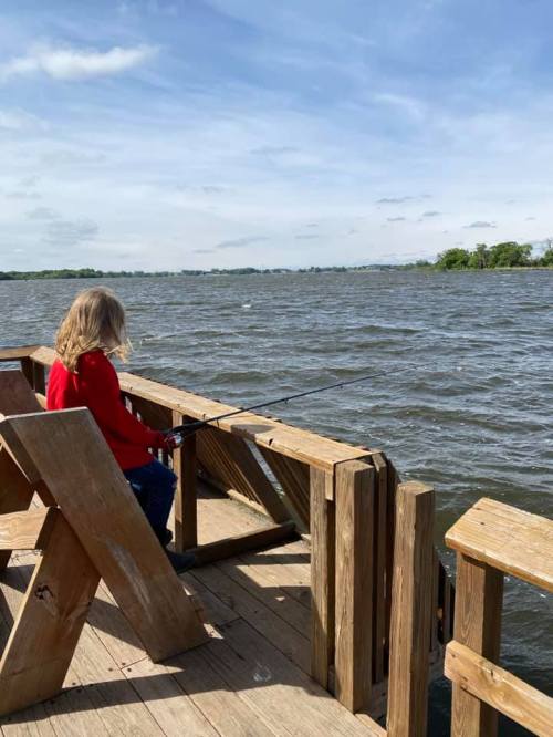 A person in a red sweater sits on a wooden dock, fishing by a lake under a blue sky with scattered clouds.