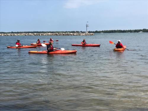 A group of people in orange kayaks paddling on a calm water body under a clear blue sky.