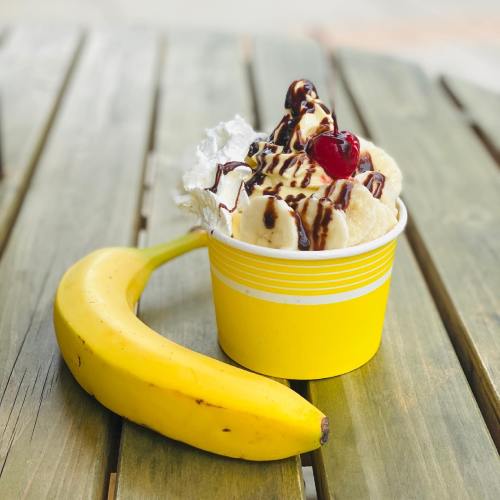 A yellow cup filled with ice cream, whipped cream, and a cherry, next to a ripe banana on a wooden table.