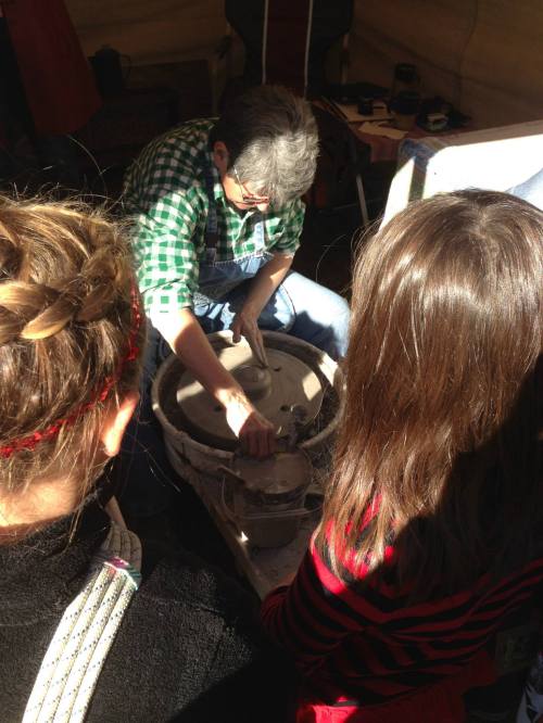 A potter shapes clay on a wheel, with two children observing intently from behind.